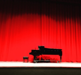 A grand piano and stool are set on a stage with bright red curtains as the backdrop. The lighting highlights the sleek black body of the piano, creating a dramatic and elegant atmosphere.