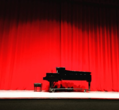 A grand piano and stool are set on a stage with bright red curtains as the backdrop. The lighting highlights the sleek black body of the piano, creating a dramatic and elegant atmosphere.
