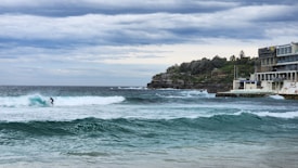 A lone surfer rides a wave in the ocean near a rocky coastline. In the background, an expansive cloudy sky stretches above, while a modern building and green foliage decorate the shoreline to the right.