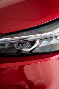 A close-up of a car's headlight against a glossy red surface, showing the intricate design of the headlight along with reflections on the surrounding bodywork.