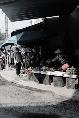 A street market scene with a variety of fruits displayed on plastic crates under a tent. Two people are visible, one standing and one crouching with a traditional conical hat, organizing the produce. The market is shaded, and the atmosphere seems busy with people in the background.