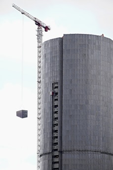 A tall construction crane is lifting a rectangular structure next to a cylindrical building that appears to be under construction. The building has a metallic or concrete texture with visible scaffolding.