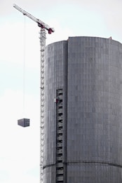 A tall construction crane is lifting a rectangular structure next to a cylindrical building that appears to be under construction. The building has a metallic or concrete texture with visible scaffolding.