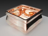 A carefully crafted glass object with square contours, featuring a metallic border and intricate swirled patterns. The interior has a molten amber appearance, reflecting light with a rich, warm glow. Resting on a smooth, light-colored surface, the background is a gradient of soft, dark tones.