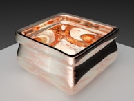 A carefully crafted glass object with square contours, featuring a metallic border and intricate swirled patterns. The interior has a molten amber appearance, reflecting light with a rich, warm glow. Resting on a smooth, light-colored surface, the background is a gradient of soft, dark tones.