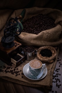 An ornate coffee grinder sits next to a cup of coffee on a decorative saucer. Coffee beans spill out of a burlap sack, scattered around a wooden surface. A smaller metal container holds additional coffee beans and a small basket contains ground coffee.