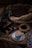 An ornate coffee grinder sits next to a cup of coffee on a decorative saucer. Coffee beans spill out of a burlap sack, scattered around a wooden surface. A smaller metal container holds additional coffee beans and a small basket contains ground coffee.