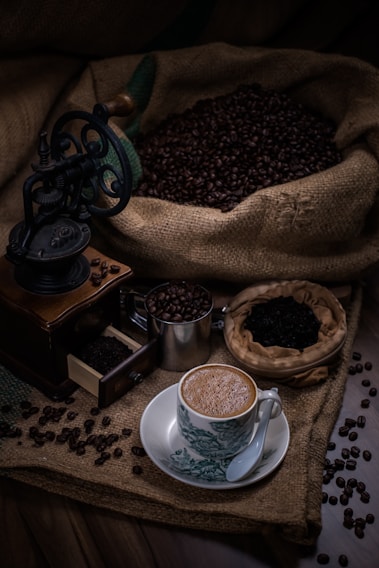 An ornate coffee grinder sits next to a cup of coffee on a decorative saucer. Coffee beans spill out of a burlap sack, scattered around a wooden surface. A smaller metal container holds additional coffee beans and a small basket contains ground coffee.