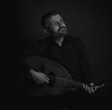 A man with a contemplative expression plays a stringed instrument, likely an oud, in a dimly lit setting. He is dressed in dark clothing, and the focus is on his thoughtful gaze, which is directed upwards.