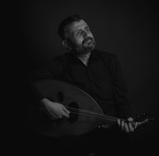 A man with a contemplative expression plays a stringed instrument, likely an oud, in a dimly lit setting. He is dressed in dark clothing, and the focus is on his thoughtful gaze, which is directed upwards.
