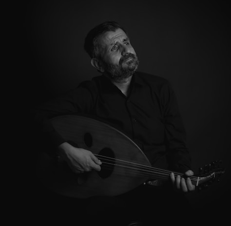 A man with a contemplative expression plays a stringed instrument, likely an oud, in a dimly lit setting. He is dressed in dark clothing, and the focus is on his thoughtful gaze, which is directed upwards.