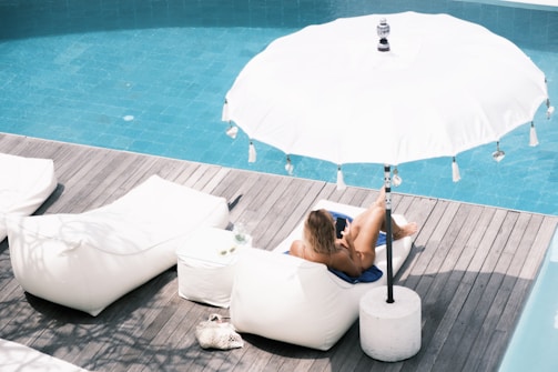 A person relaxes on a white lounger under a large white beach umbrella beside a blue swimming pool. The scene is set on a wooden deck with a small white side table and a woven bag nearby. The person is holding a black smartphone and appears to be enjoying the sun.
