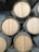 Several wooden barrels are stacked in a storage facility, with the ends facing outward. The barrels are cylindrical with metal hoops securing the wooden slats. The surrounding environment includes corrugated metal walls and a dimly lit atmosphere.