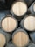 Several wooden barrels are stacked in a storage facility, with the ends facing outward. The barrels are cylindrical with metal hoops securing the wooden slats. The surrounding environment includes corrugated metal walls and a dimly lit atmosphere.