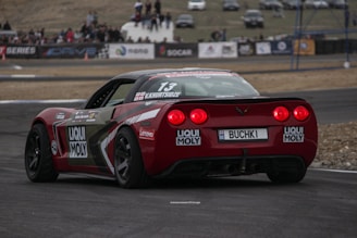 A red racing car is on a track with branding decals, including Liqui Moly and Lenovo. The car has a sleek design with a spoiler and visible track conditions suggest high-speed racing. In the background, spectators are gathered around the track, observing the event.