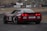 A red racing car is on a track with branding decals, including Liqui Moly and Lenovo. The car has a sleek design with a spoiler and visible track conditions suggest high-speed racing. In the background, spectators are gathered around the track, observing the event.