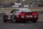 A red racing car is on a track with branding decals, including Liqui Moly and Lenovo. The car has a sleek design with a spoiler and visible track conditions suggest high-speed racing. In the background, spectators are gathered around the track, observing the event.