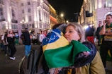 A woman is embracing someone while holding a Brazilian flag in a lively, well-lit urban area at night. Several people are in the background, some wearing casual clothes and others holding flags, creating a bustling atmosphere.