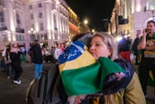 A woman is embracing someone while holding a Brazilian flag in a lively, well-lit urban area at night. Several people are in the background, some wearing casual clothes and others holding flags, creating a bustling atmosphere.