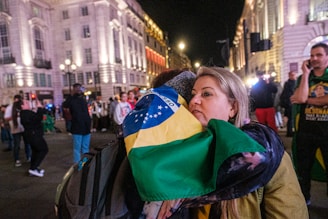 A woman is embracing someone while holding a Brazilian flag in a lively, well-lit urban area at night. Several people are in the background, some wearing casual clothes and others holding flags, creating a bustling atmosphere.