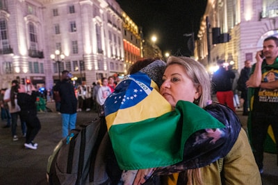 A woman is embracing someone while holding a Brazilian flag in a lively, well-lit urban area at night. Several people are in the background, some wearing casual clothes and others holding flags, creating a bustling atmosphere.