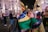 A woman is embracing someone while holding a Brazilian flag in a lively, well-lit urban area at night. Several people are in the background, some wearing casual clothes and others holding flags, creating a bustling atmosphere.