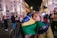 A woman is embracing someone while holding a Brazilian flag in a lively, well-lit urban area at night. Several people are in the background, some wearing casual clothes and others holding flags, creating a bustling atmosphere.