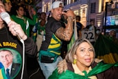 A group of people is gathered, one holding a megaphone and others wearing green, yellow, and blue clothing, indicative of support for Brazil. Flags, banners, and patriotic colors are prominent amid an urban nighttime setting. One person is singing or speaking passionately with a microphone.