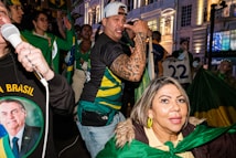 A group of people is gathered, one holding a megaphone and others wearing green, yellow, and blue clothing, indicative of support for Brazil. Flags, banners, and patriotic colors are prominent amid an urban nighttime setting. One person is singing or speaking passionately with a microphone.