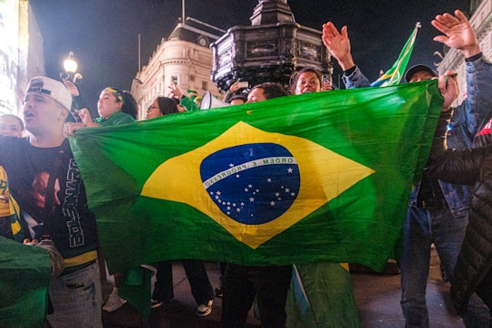 A group of people are gathered at night, holding a large Brazilian flag. The background features an impressive historic building and street lamps, indicating an urban setting. The crowd appears engaged and possibly celebrating, with people raising their arms or gesturing animatedly.