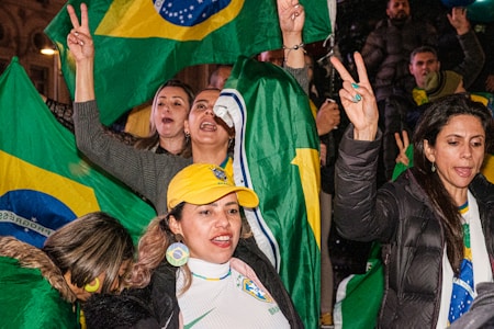 A group of enthusiastic people, predominantly women, are celebrating. They are holding and waving flags with a green, yellow, and blue design. Some are making peace signs with their fingers, and one woman is wearing a yellow cap with a matching flag design on her shirt. The scene conveys a sense of unity and excitement.