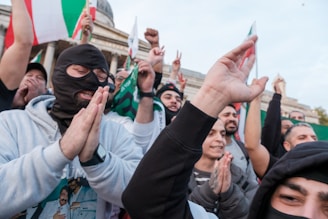 A group of people gathered, appearing to be in a celebratory or protest setting. Some are cheering, with one individual wearing a mask and others clapping or raising their hands. A background building with columns and flags held by participants suggests a public or political event.