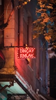 A glowing red neon sign with the words 'Dinçay Emlak' is prominently displayed on the side of a building. The surface of the wall is textured with stone, and surrounding the sign are hanging cables. Leafy branches from a nearby tree cascade in from the top right, casting shadows on the wall, and contributing to a mix of warm and cool tones, creating a sense of depth.