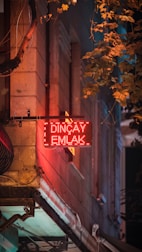 A glowing red neon sign with the words 'Dinçay Emlak' is prominently displayed on the side of a building. The surface of the wall is textured with stone, and surrounding the sign are hanging cables. Leafy branches from a nearby tree cascade in from the top right, casting shadows on the wall, and contributing to a mix of warm and cool tones, creating a sense of depth.