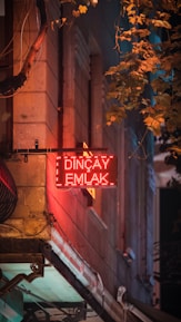 A glowing red neon sign with the words 'Dinçay Emlak' is prominently displayed on the side of a building. The surface of the wall is textured with stone, and surrounding the sign are hanging cables. Leafy branches from a nearby tree cascade in from the top right, casting shadows on the wall, and contributing to a mix of warm and cool tones, creating a sense of depth.