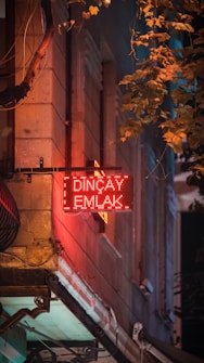 A glowing red neon sign with the words 'Dinçay Emlak' is prominently displayed on the side of a building. The surface of the wall is textured with stone, and surrounding the sign are hanging cables. Leafy branches from a nearby tree cascade in from the top right, casting shadows on the wall, and contributing to a mix of warm and cool tones, creating a sense of depth.