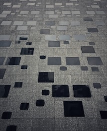 A building facade composed of numerous square and rectangular windows, varying in size. The surface has a textured, metallic appearance with a gradient of dark to light shades. The layout of the windows creates an abstract and modern aesthetic.