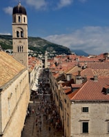 A scenic view of a historic city street with a tall bell tower to the left. The street is lined with old buildings featuring red-tiled roofs, and a crowd of people is visible below, walking along the paved pathway. Hills covered in greenery form the backdrop under a partially cloudy sky.