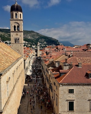 A scenic view of a historic city street with a tall bell tower to the left. The street is lined with old buildings featuring red-tiled roofs, and a crowd of people is visible below, walking along the paved pathway. Hills covered in greenery form the backdrop under a partially cloudy sky.
