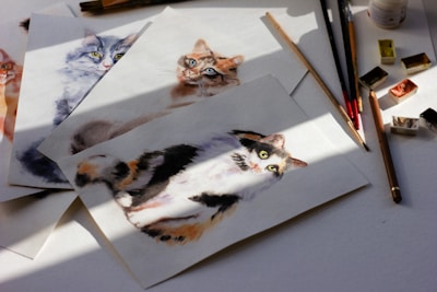 A collection of watercolor paintings depicting different cats is arranged on a surface. The paintings feature various cat breeds and poses, characterized by soft brush strokes and vibrant colors. Paintbrushes and paint pans are scattered around the artwork, suggesting an artistic work in progress under natural light, casting soft shadows.