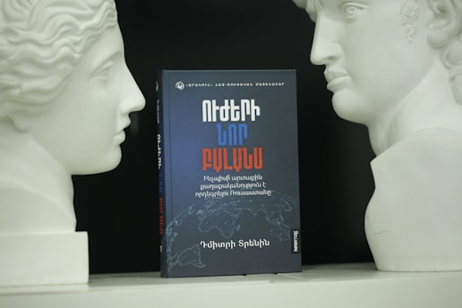 A book with a black and red cover, displaying text in a non-Latin script, is positioned between two white bust sculptures facing each other. The background is dark, highlighting the objects in the foreground.