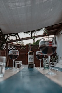 A rustic outdoor setting featuring a long, narrow table with a bottle of red wine in the center, flanked by three glasses partially filled with red wine. The scene is set under a canopy, with some foliage visible in the background.