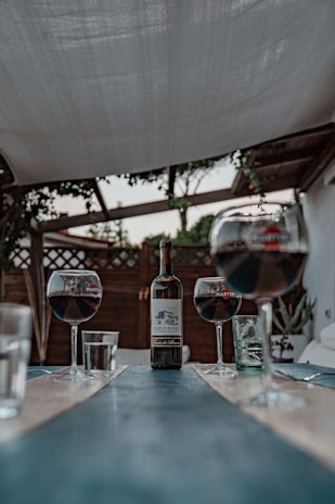 A rustic outdoor setting featuring a long, narrow table with a bottle of red wine in the center, flanked by three glasses partially filled with red wine. The scene is set under a canopy, with some foliage visible in the background.