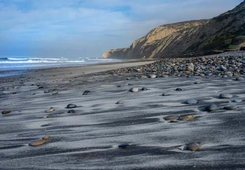 A serene rocky beach with smooth black and gray stones under a bright blue sky dotted with fluffy white clouds.