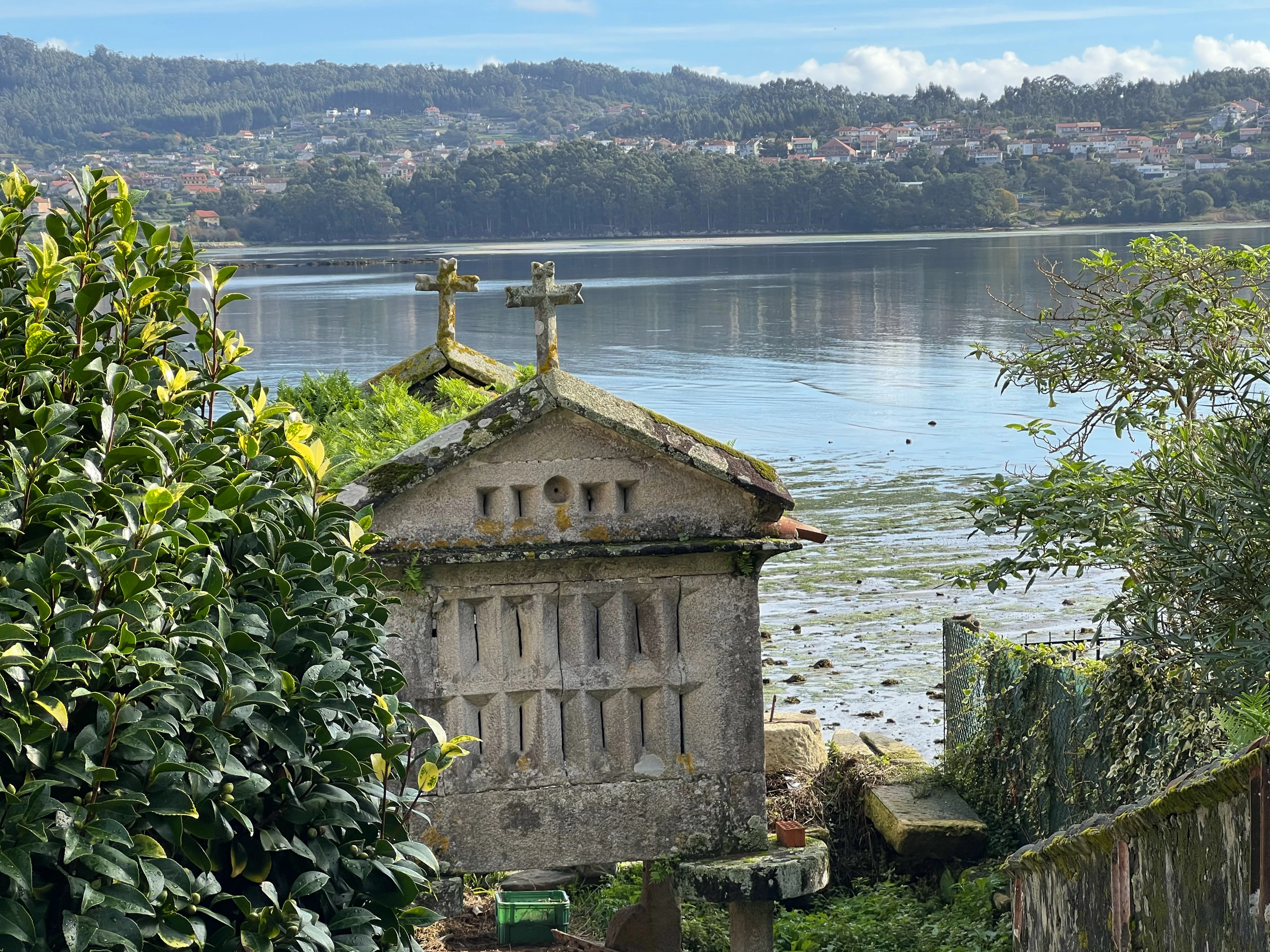 A stone hórreo topped with crosses beside a calm Galician inlet