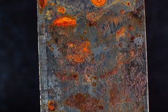 A close-up view of a rusted metal surface displaying various shades of orange and brown rust spots. The texture appears rough and uneven, with patches of corrosion scattered across the surface.