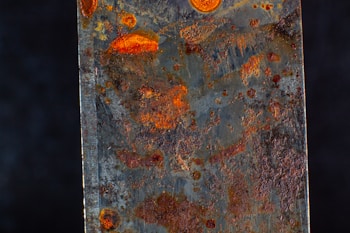 A close-up view of a rusted metal surface displaying various shades of orange and brown rust spots. The texture appears rough and uneven, with patches of corrosion scattered across the surface.