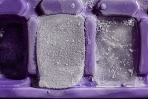 An extreme close-up of a purple ice cube tray with partially frozen ice cubes. The ice cubes have varying levels of frost and ice crystallization on them.