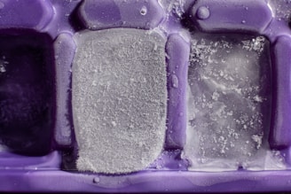 An extreme close-up of a purple ice cube tray with partially frozen ice cubes. The ice cubes have varying levels of frost and ice crystallization on them.