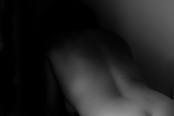 A black and white photograph featuring a person's back with a focus on the curve of the spine. The lighting is dim, creating a shadowy, mysterious effect.